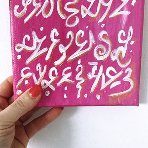 Op de afbeelding: Een roze canvas schilderij met witte en gouden abstracte kalligrafie. Het schilderij is op een witte achtergrond.