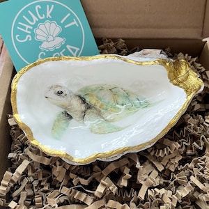 May include: A decorative oyster shell featuring a hand-painted sea turtle on the interior with a gold-painted rim. The shell is nestled in a cardboard box with brown paper filler and includes a teal square card with the text Shuck It And Sea.