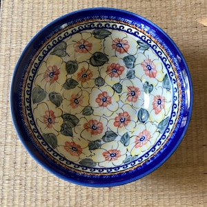 May include: A round ceramic bowl with a vibrant floral pattern. The bowl features a deep blue rim and a decorative border. The interior is adorned with a repeating pattern of white flowers with orange centers and green leaves, set against a yellow background.