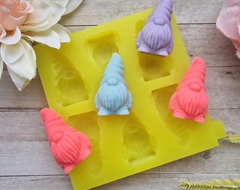 May include: A yellow silicone mold with six gnome-shaped cavities. Three finished gnomes in pastel colors: pink, blue, and purple, are displayed. The mold and gnomes are on a light-colored surface with floral accents.