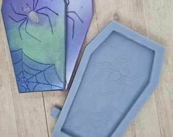 May include: A gray silicone mold shaped like a coffin with a spider and web design. The mold is used to create resin crafts.