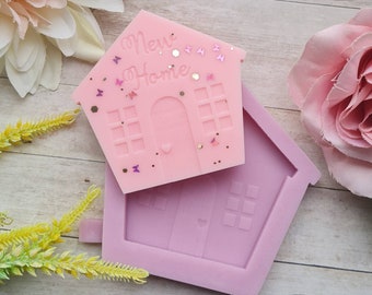 May include: Pink silicone mould shaped like a house with the words "New Home" on the top. The mould is open and a pink house-shaped candle is sitting on top of it.