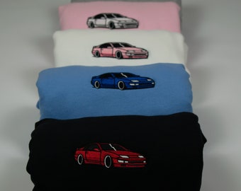 May include: A stack of sweatshirts in various colors, each featuring an embroidered image of a red, blue, white, or pink car. The sweatshirts are folded and stacked on top of each other.