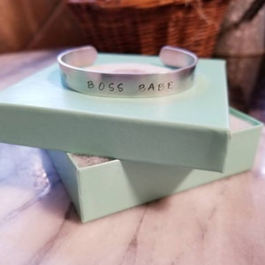 May include: A silver metal bracelet with the words "Boss Babe" engraved on it. The bracelet is in a light blue gift box.
