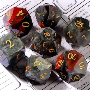 May include: A set of seven polyhedral dice with a faceted, semi-translucent appearance. The dice feature a mix of deep red and mossy green inclusions, with engraved gold-colored numbers. The dice are arranged on a white paper game sheet.