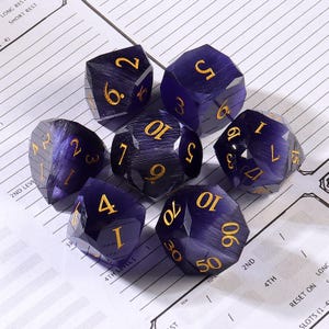 May include: Set of seven faceted, purple dice with gold numbering. The dice are arranged on a white surface with printed text and lines, suitable for tabletop gaming. Each die displays different numbers.