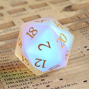 May include: A frosted, iridescent twenty-sided die with gold-colored numerals. The die is resting on a background of a game sheet. The numbers are clearly visible, with the number 20 prominently displayed.