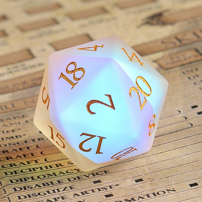 May include: A frosted, iridescent twenty-sided die with gold-colored numerals. The die is resting on a background of a game sheet. The numbers are clearly visible, with the number 20 prominently displayed.