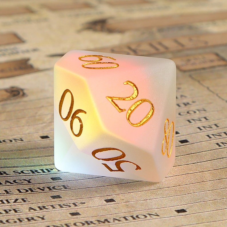 May include: A frosted, multi-sided die with gold-colored numbers. The die has a gradient of colors, transitioning from yellow to pink. The numbers visible are 06, 20, and 50. The die is likely used for tabletop games.