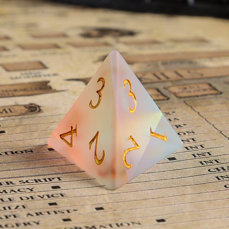 May include: A translucent pyramid-shaped die with gold-colored numerals. The die has a gradient of colors, transitioning from peach to white. The numbers 1, 2, 3, and 4 are visible. The die is likely for tabletop games.