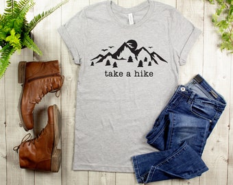 May include: A gray t-shirt with a black mountain range graphic and the text "take a hike". The shirt is laid out on a wooden surface with a pair of brown leather boots and blue jeans.