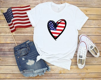 Può includere: Maglietta bianca con un design a forma di cuore rosso, bianco e blu con la bandiera americana. La maglietta è abbinata a pantaloncini di jeans e scarpe da ginnastica bianche.