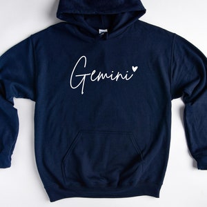May include: A navy blue hoodie with a white cursive inscription that reads "Gemini" with a small white heart to the right of the word.