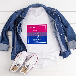 Könnte beinhalten: Weißes T-Shirt mit einem Grafikdruck eines rosa, lila und blauen Quadrats mit den Buchstaben "BI" in Weiß und dem Text "Bisexuell 83" darunter. Das T-Shirt liegt auf einer weißen Holzoberfläche mit einer blauen Jeansjacke und weißen Turnschuhen.