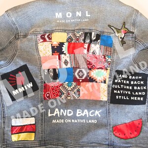 May include: A light blue denim jacket with a patchwork design on the back. The patches are made of different fabrics and feature various colors and patterns. The jacket also has several embroidered patches, including a hummingbird, a watermelon, and a handprint with the text "MMIW". The text "MONL MADE ON NATIVE LAND" is printed on the back of the jacket. The text "LAND BACK MADE ON NATIVE LAND" is also printed on the back of the jacket. The text "LAND BACK WATER BACK CULTURE BACK NATIVE LAND STILL HERE" is printed on a black patch on the back of the jacket.