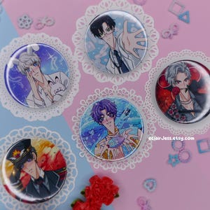 Puede incluir: Cinco botones redondos de personajes de estilo anime. Cada botón presenta un diseño de personaje diferente con colores vibrantes e ilustraciones detalladas. Los botones están colocados sobre tapetes de encaje blanco decorativos sobre un fondo rosa y azul.