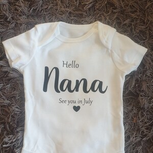 以下が含まれることがあります： 黒い文字で「Hello Nana See you in July」と書いてある白いベビーロンパース。小さな黒いハート付き。