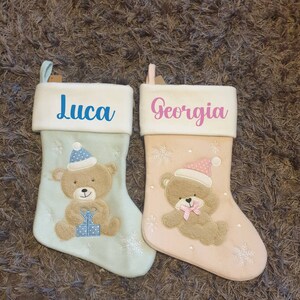 Può includere: Due calze di Natale personalizzate con orsacchiotti ricamati e fiocchi di neve. La calza a sinistra è azzurra con il nome "Luca" ricamato. La calza a destra è rosa chiaro con il nome "Georgia" ricamato.