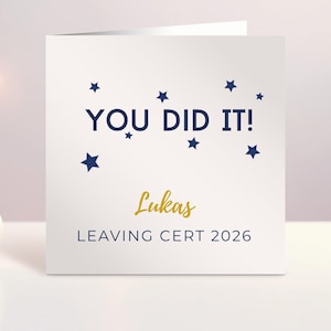 Może przedstawiać: Kwadratowa biała kartka okolicznościowa z ciemnoniebieskim napisem YOU DID IT! otoczonym małymi ciemnoniebieskimi gwiazdkami. Poniżej znajduje się imię Lukas wydrukowane złotą kursywą, a na dole ciemnoniebieski napis LEAVING CERT 2026.