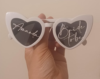 May include: White heart-shaped sunglasses with dark lenses. The left lens features the white script text Amanda, and the right lens features the white script text Bride to be. The frames are made of a solid white plastic material with a cat-eye shape.