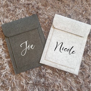 Può includere: Due custodie per laptop in feltro, una grigia e una bianca, con i nomi "Joe" e "Nicole" scritti in lettere bianche e nere, rispettivamente.