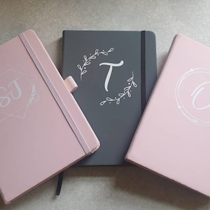 Puede incluir: Tres cuadernos personalizados con diferentes iniciales y diseños. Los cuadernos son rosas y grises con letras blancas. Las iniciales son "SJ", "T" y "O".
