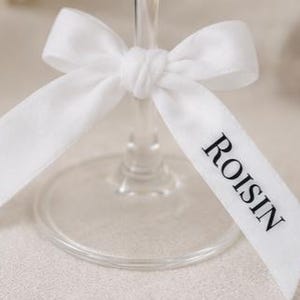 May include: A white satin ribbon tied in a bow around the stem of a clear glass. The ribbon features the name ROISIN printed in black capital letters on one of the trailing ends. The glass has a circular base visible beneath the tied ribbon.
