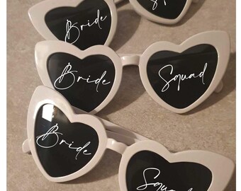 May include: Three pairs of white heart-shaped sunglasses with dark lenses. The left lens of each pair features the white script text "Bride" and the right lens features the white script text "Squad" printed across the surface of the lenses.