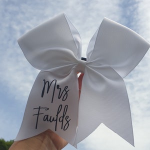 May include: White hair bow with a black script that reads "Mrs. Faulds".