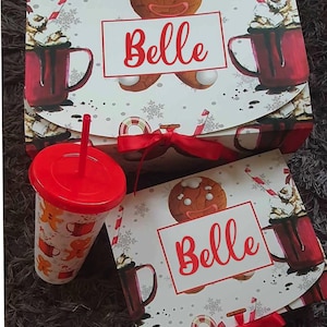 Könnte beinhalten: Eine weiße Geschenkbox und ein passender Kunststoffbecher mit rotem Deckel und Strohhalm. Die Box und der Becher zeigen ein festliches Muster aus Lebkuchenmännern, roten Tassen mit heißer Schokolade und Schneeflocken. Der Name Belle steht in Rot.