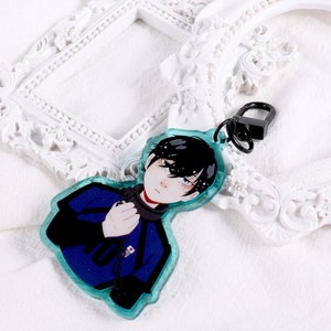 Peut inclure: Un porte-clés représentant un personnage d'anime bleu et noir sur fond blanc. Le porte-clés est attaché à une chaîne argentée et pend à un cadre blanc orné.