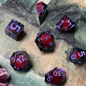 Puede incluir: Conjunto de siete dados rojos y morados con números blancos. Los dados están sobre una superficie cuadriculada con un diseño de mapa.