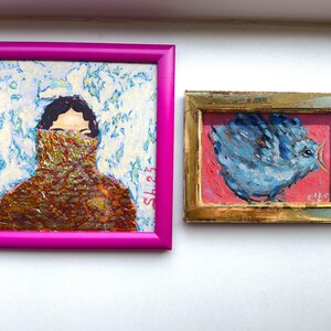 Pode incluir: Duas pinturas emolduradas. A maior, em uma moldura magenta, retrata uma pessoa com uma roupa estampada cobrindo a parte inferior do rosto. A pintura menor, em uma moldura dourada, mostra um p&aacute;ssaro azul em um fundo rosa.
