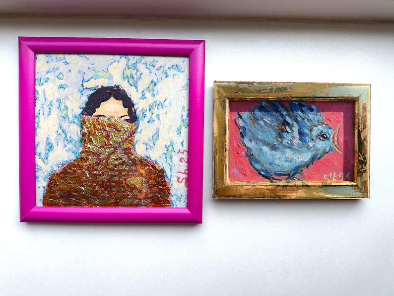 Pode incluir: Duas pinturas emolduradas. A maior, em uma moldura magenta, retrata uma pessoa com uma roupa estampada cobrindo a parte inferior do rosto. A pintura menor, em uma moldura dourada, mostra um p&aacute;ssaro azul em um fundo rosa.