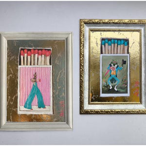 Pode incluir: Duas obras de arte emolduradas com designs de caixas de f&oacute;sforos. Uma mostra uma figura com cal&ccedil;as turquesa e um fundo rosa, a outra um sapo com um fato azul. Ambas t&ecirc;m f&oacute;sforos no topo e est&atilde;o em molduras decorativas.