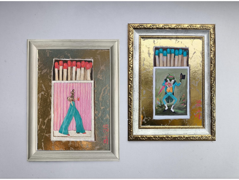 Pode incluir: Duas obras de arte emolduradas com designs de caixas de f&oacute;sforos. Uma mostra uma figura com cal&ccedil;as turquesa e um fundo rosa, a outra um sapo com um fato azul. Ambas t&ecirc;m f&oacute;sforos no topo e est&atilde;o em molduras decorativas.