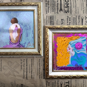 Pode incluir: Duas pinturas emolduradas com molduras douradas e brancas ornamentadas. A pintura da esquerda retrata uma figura em um vestido roxo com um len&ccedil;o branco. A pintura da direita mostra uma mulher com cabelo amarelo e rosto azul. Ambas as pinturas s&atilde;o assinadas.