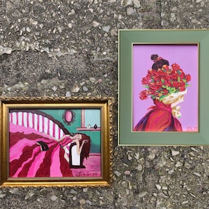 Pode incluir: Duas pinturas emolduradas. Uma apresenta uma mulher com um vestido rosa reclinada em uma cama listrada. A outra retrata uma figura com uma cabe&ccedil;a de flores vermelhas em um fundo roxo. Ambas s&atilde;o exibidas em uma superf&iacute;cie cinza texturizada.