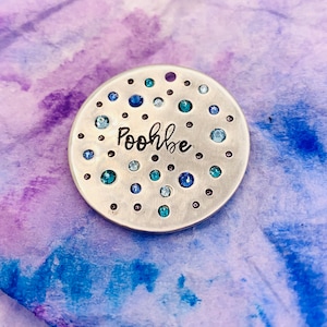 May include: A silver round tag with a scattered pattern of blue and teal rhinestones. The tag is engraved with the name "Poohbe".