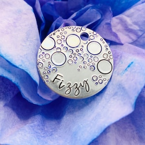 May include: Silver charm with a bubble design and the word "Fizzyg" engraved on it. The charm is on a blue and purple background.