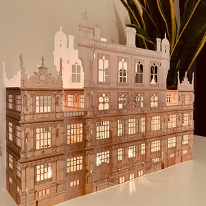 Peut inclure: Un modèle en papier d'un grand bâtiment orné avec de nombreuses fenêtres et une façade détaillée. Le modèle est éclairé de l'intérieur, créant une lueur chaleureuse.