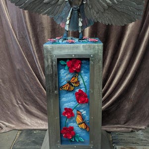 May include: A metal sculpture of a bird with outstretched wings atop a rectangular pedestal. The pedestal features a blue background with red roses and orange butterflies. The sculpture is made of various metal pieces.