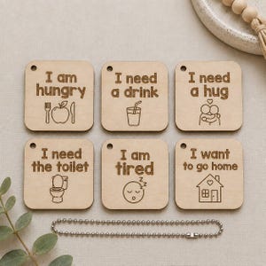 May include: Six square light-coloured wooden tags with rounded corners, each featuring a small hole at the top left. The tags are engraved with text and icons: I am hungry, I need a drink, I need a hug, I need the toilet, I am tired, and I want to go home. A silver metal ball chain is included.