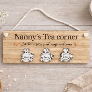 May include: A wooden sign with the text "Nanny's Tea corner" and "Little visitors always welcome." It has three teacup designs with names "Woody," "Jakey," and "Lori." A rope is attached for hanging. The sign is made of light-coloured wood.