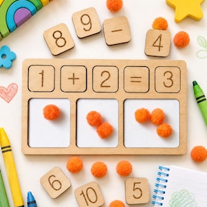 May include: A wooden maths learning board with square tiles featuring numbers 1, 2, 3, 5, 6, 8, 9, 10 and symbols plus, minus, and equals. The board has three white rectangular slots containing orange pom-poms, accompanied by crayons and a notebook.