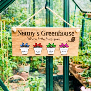 May include: Wooden sign reading "Nanny's Greenhouse" and "Where little loves grow..." with four flower pots labelled Harry, Leo, Albie, and Ava. The sign is suspended in a greenhouse, with plants visible in the background.