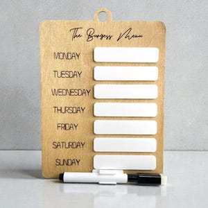 May include: A wooden weekly menu board, labelled "The Burgess Menu". It features seven white rectangular spaces for each day of the week, and a black and white marker. The board is made of light brown wood.