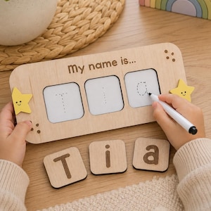 May include: A rectangular wooden educational board with the text My name is... featuring three white square inserts with dotted letters for tracing. The board has yellow star accents on the sides and comes with three separate wooden tiles displaying the letters T, i, and a.