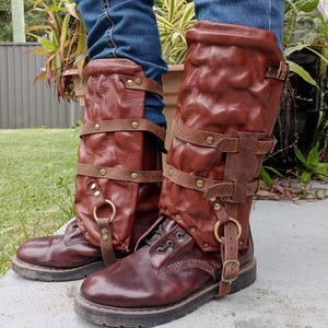 May include: Pair of burgundy leather boots with brown leather leg wraps. The boots have a lace-up design and a thick sole. The leg wraps are secured with straps and buckles, and feature brass-colored metal rings.