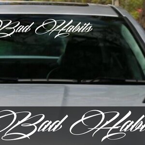 May include: White vinyl decal with the words "Bad Habits" in a cursive font. The decal is displayed on a car windscreen. A second decal is shown below the windscreen.